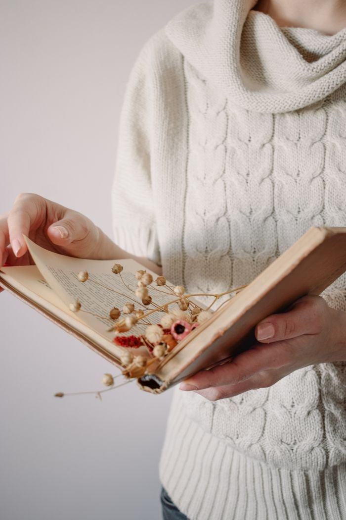 A woman wearing a white sweater holds an open vintage book with dried flowers between the pages.