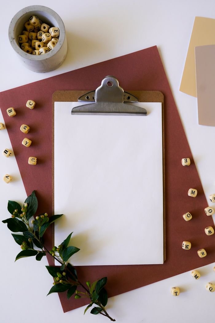 Flatlay of a clipboard with letter blocks and greenery on a white surface.
